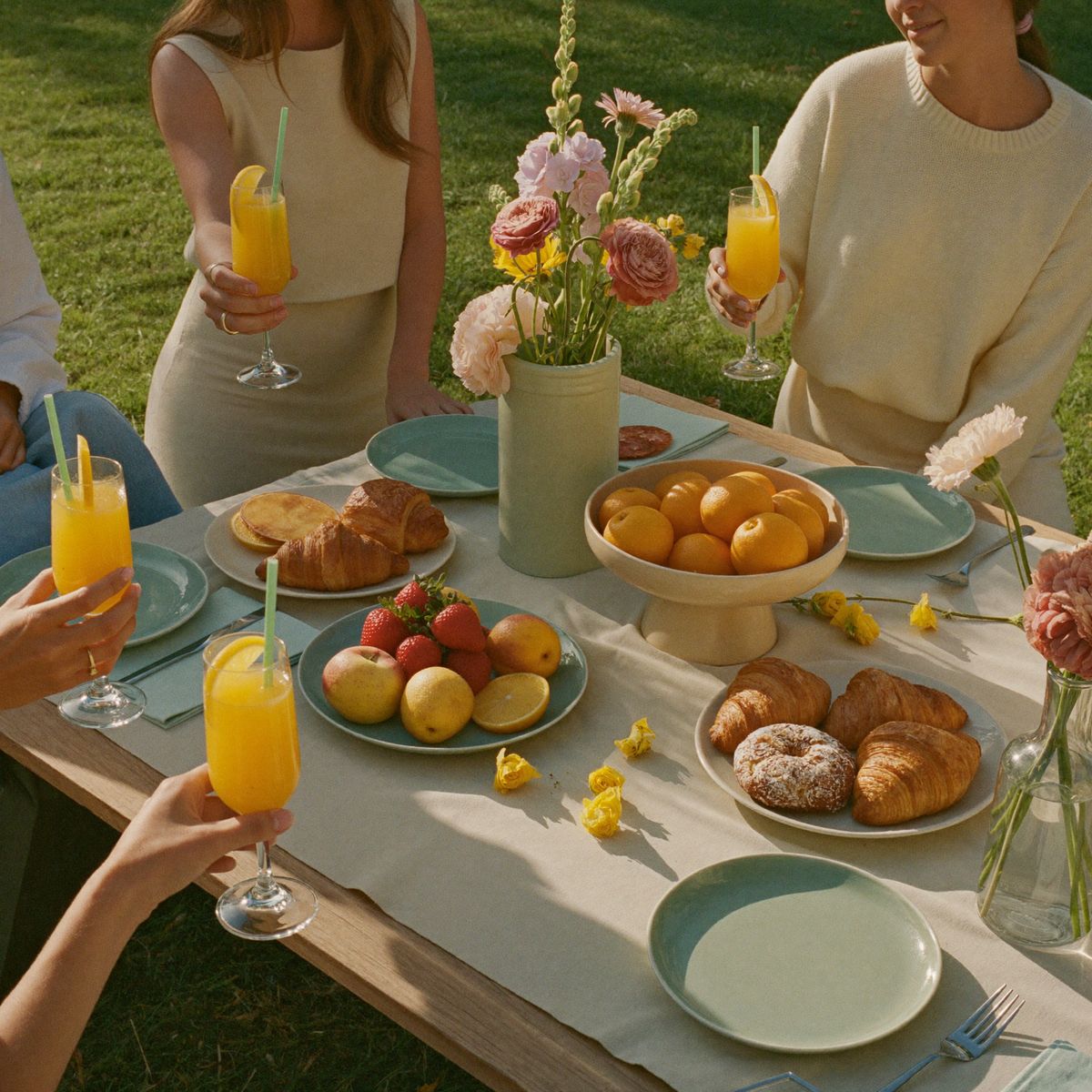 Friends raising mimosas over a styled brunch picnic table with croissants and fresh fruit