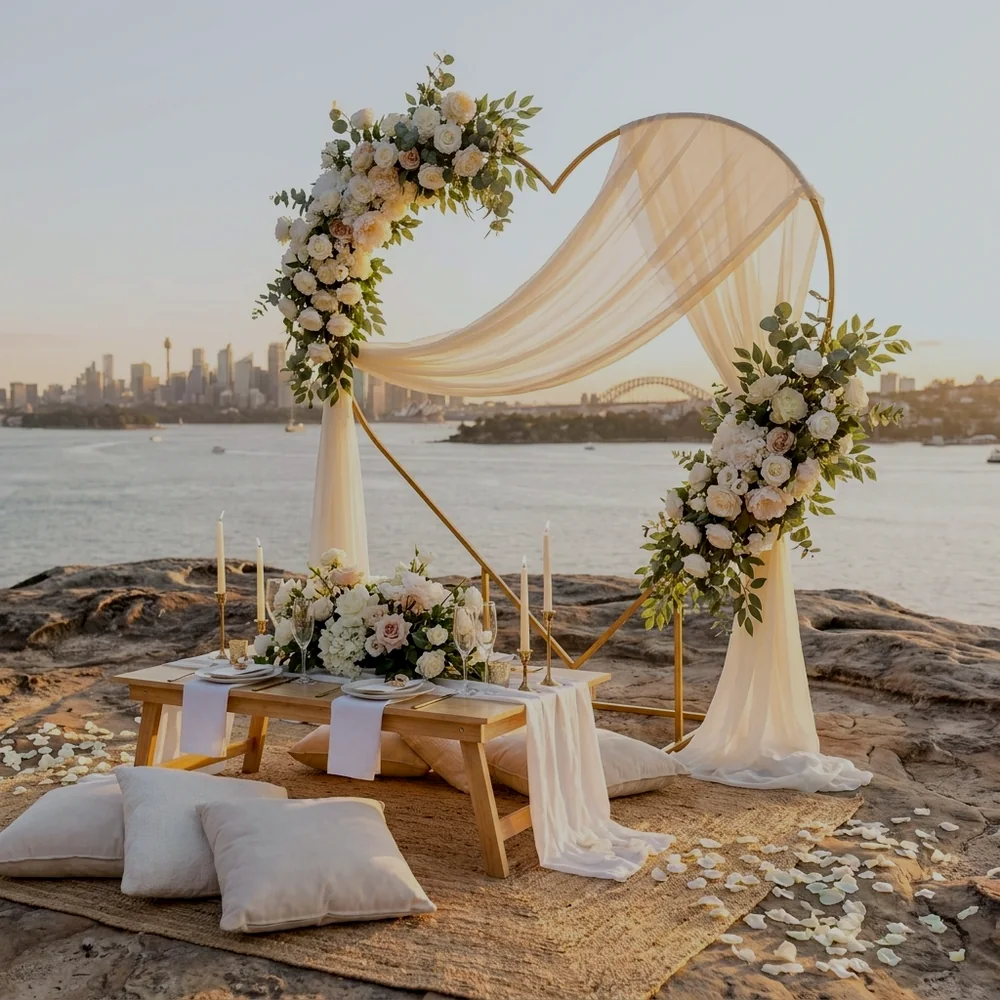 All-white proposal setup with a floral heart arch, ivory drapes and a low timber table above Sydney Harbour