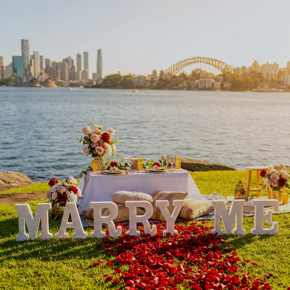 Proposal setup with large white MARRY ME letters and a red rose petal pathway on a harbourside lawn