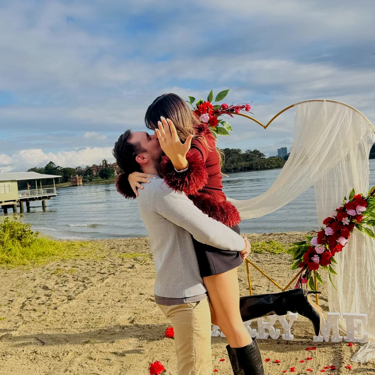 A newly engaged couple celebrating beside the floral heart arch Harmony Moments styled on a Sydney beach
